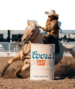 A barrel racer in a white hat turns her palomino tight around a Coors Banquet barrel.