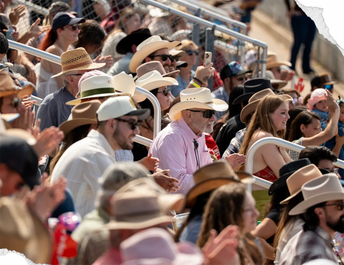 A packed grandstand of rodeo fans in cowboy hats, some of them clapping.