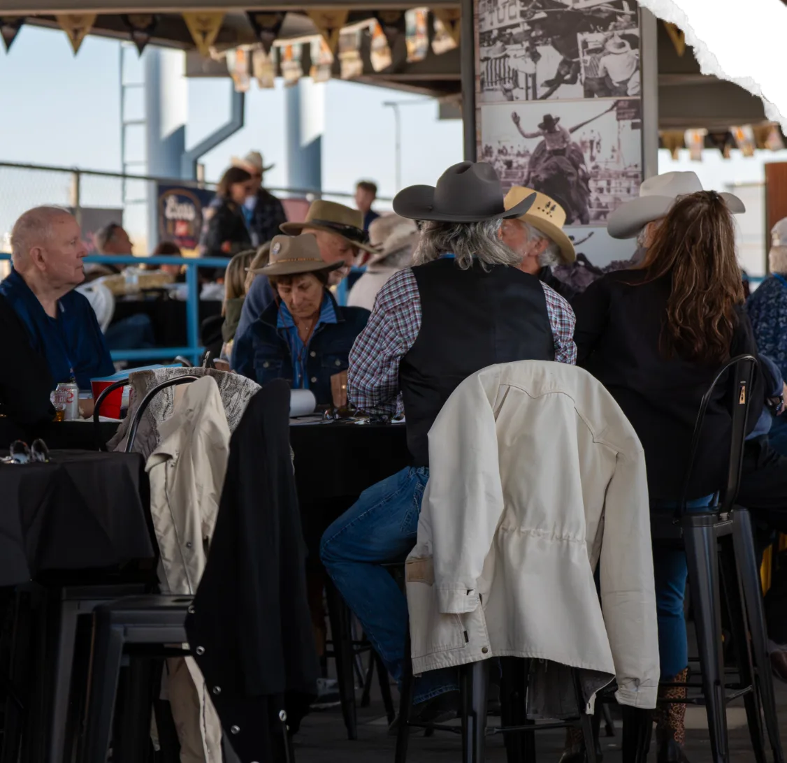 Guests in cowboy hats sit at tables in a covered hospitality area, jackets hung over their chairs.